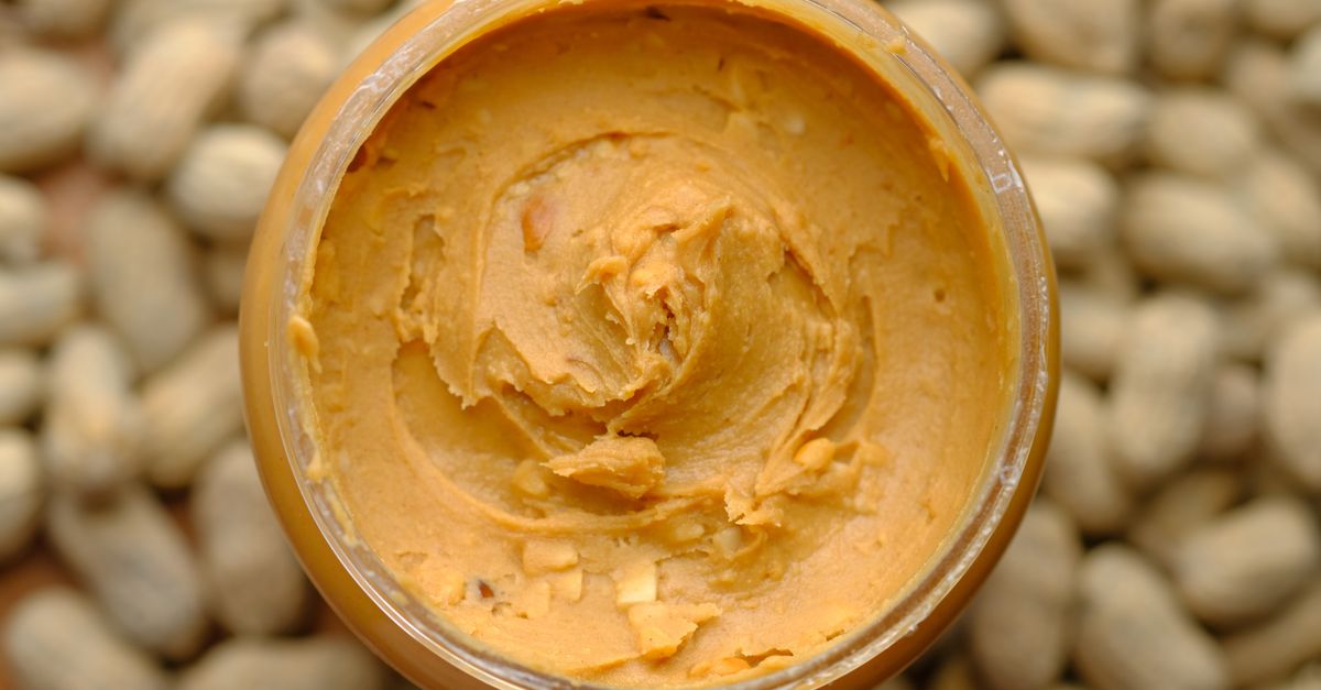 Close-up of creamy peanut butter in a glass jar surrounded by peanuts. Tasty and homemade.